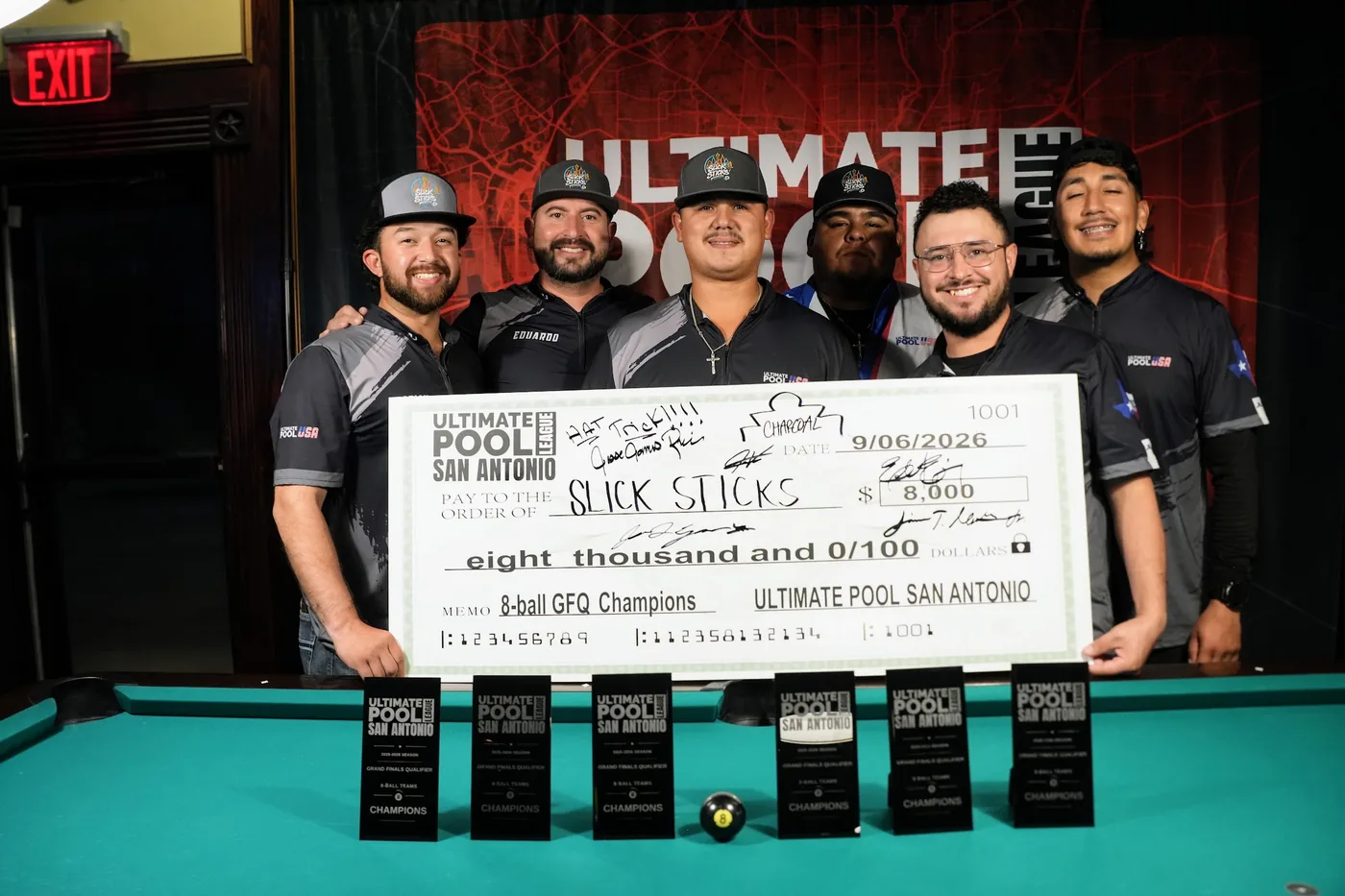 Slick Sticks holding the $8,000 travel-fund check dated 9/06/2026, made out to them as 8-Ball GFQ Champions, in front of the Ultimate Pool League backdrop at Fast Eddie's Braun with the row of champions' trophies on the table in front.