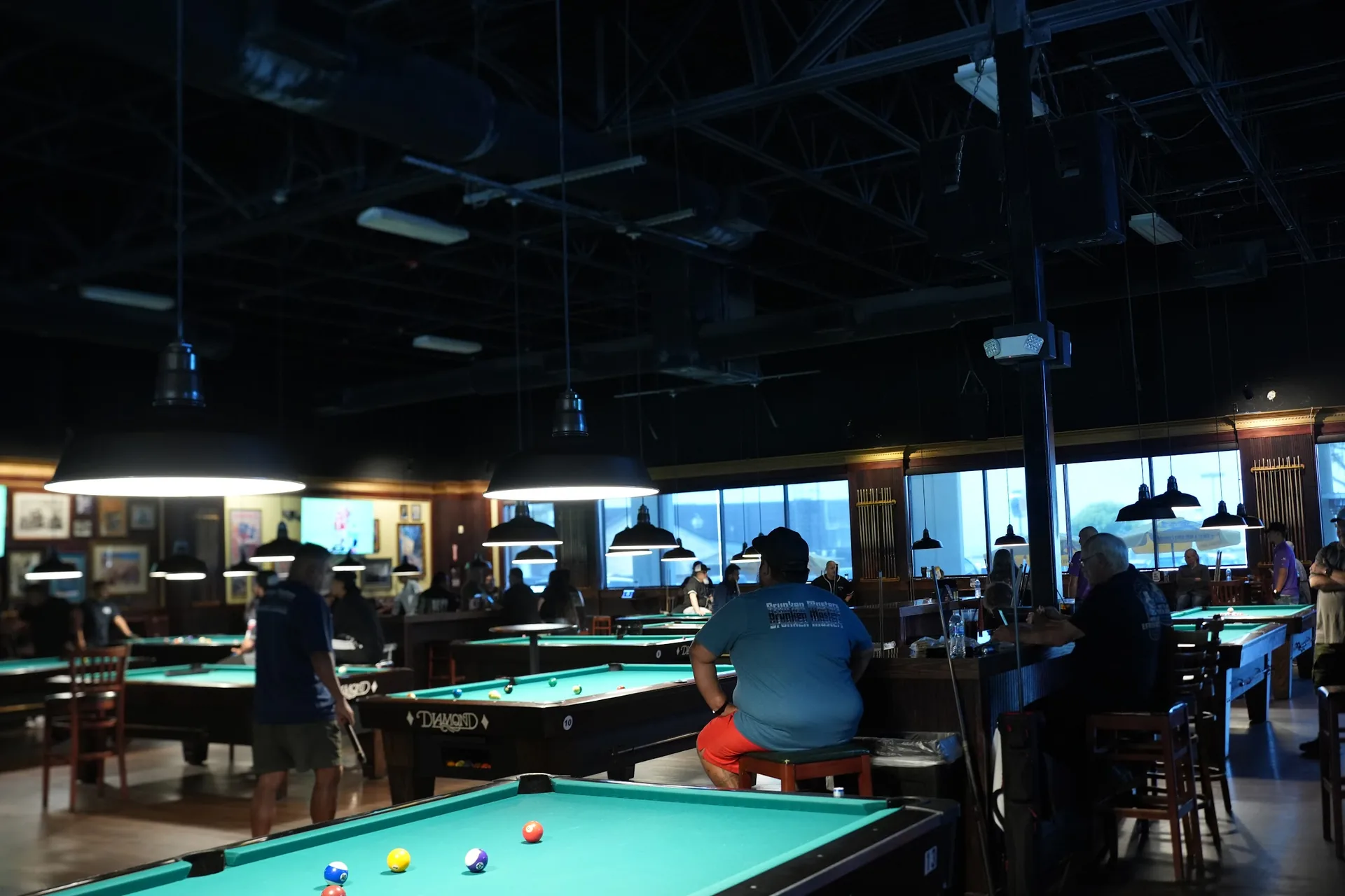 The room at Fast Eddie's Braun on the Saturday of the 8-Ball qualifier: tables in play under the pendant lights, players and spectators along the rail, daylight in the windows behind.